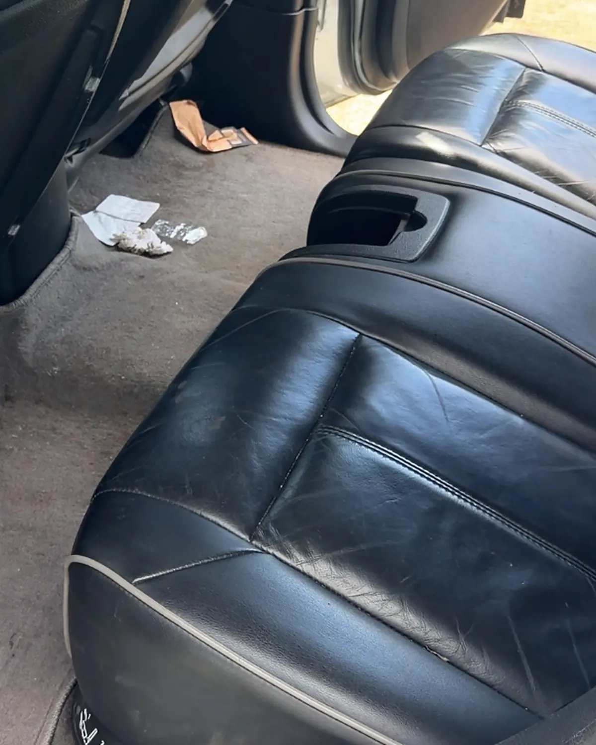 A black leather rear bench seat with paper litter and debris scattered across the grey carpet beside it